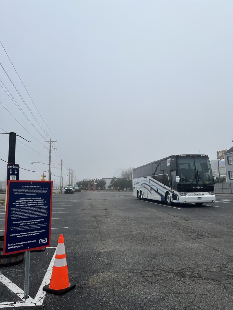A white and blue coach bus is parked in a mostly empty lot on a foggy day. An orange traffic cone and a blue PMC sign with white and red text are in the foreground near a parking meter.