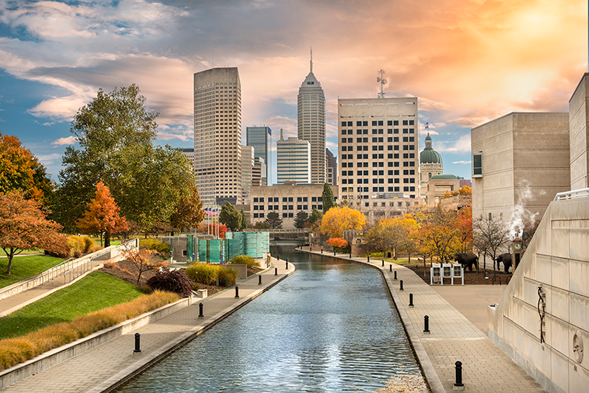 A scenic canal runs through downtown Indianapolis, flanked by walkways, autumn trees, and modern buildings under a vibrant, partly cloudy sky at sunset.