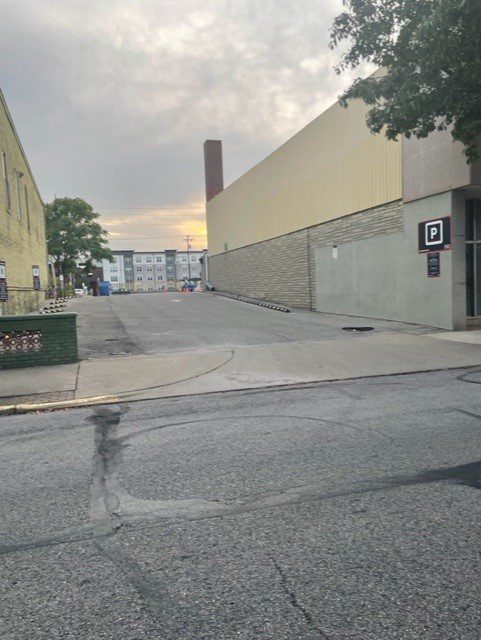 Empty urban alley with a parking lot sign on a building, concrete pavement, and a cloudy sky. In the background, there are more buildings and a chimney. A tree partially covers the top right corner.
