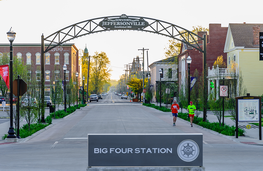 Two people jog down a wide street under an archway reading "Downtown Jeffersonville." Buildings, streetlights, and greenery line the road. A sign in the foreground reads "Big Four Station.