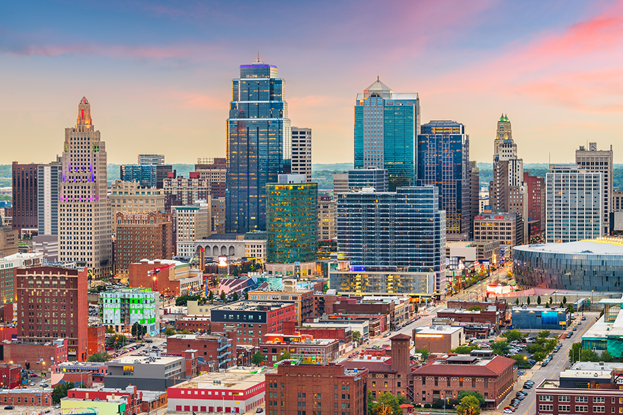 Kansas City skyline at sunset with tall skyscrapers, historic buildings, and the colorful sky in the background. The cityscape shows a mix of modern and older architecture.