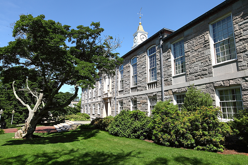 A large stone building with tall windows and a clock tower stands behind a green lawn and leafy trees on a sunny day.