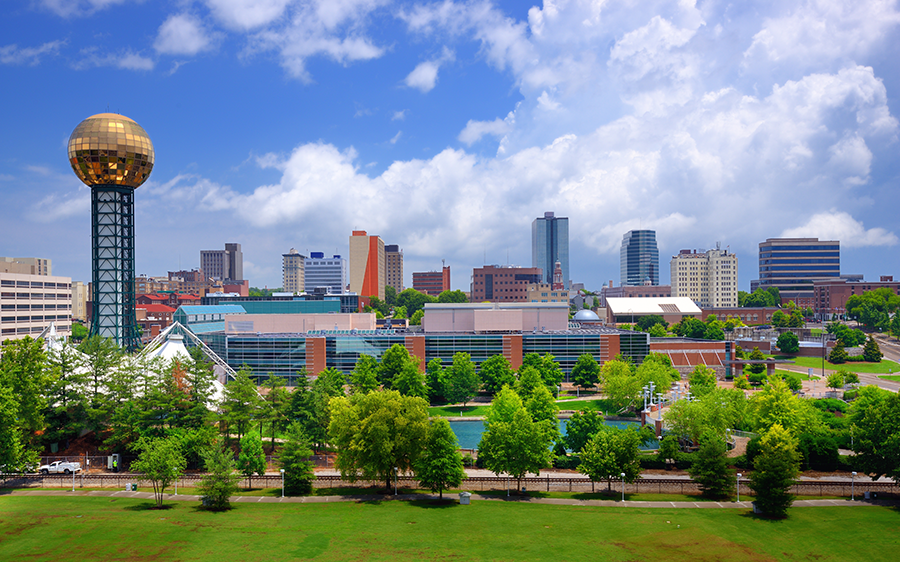 A cityscape featuring a tall structure with a golden sphere, modern buildings, green trees, and a grassy park under a partly cloudy sky.