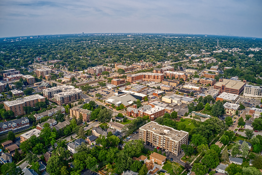 Aerial view of a suburban cityscape with residential buildings, streets lined with trees, and low-rise commercial structures under a partly cloudy sky. Dense greenery surrounds the urban area, extending to the horizon.