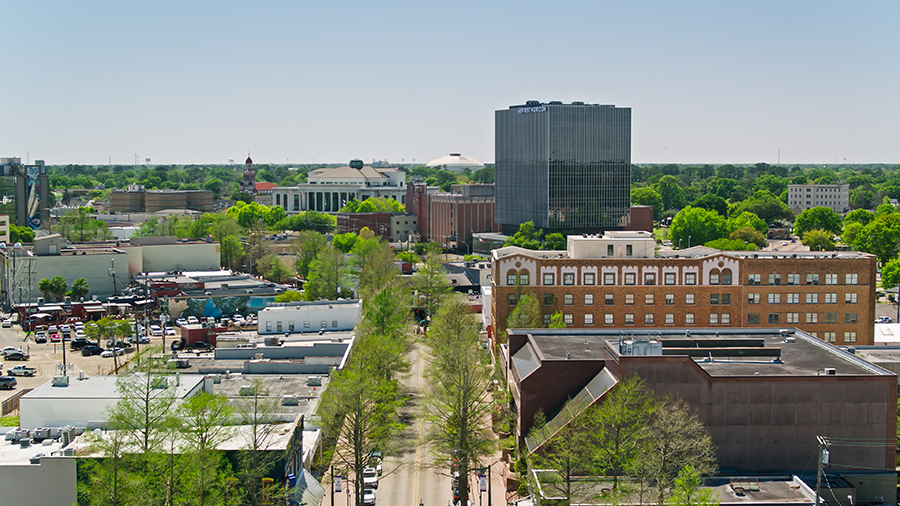 Aerial view of a small city downtown with tree-lined streets, mid-rise buildings, and a mix of older brick structures and modern glass offices under a clear blue sky.