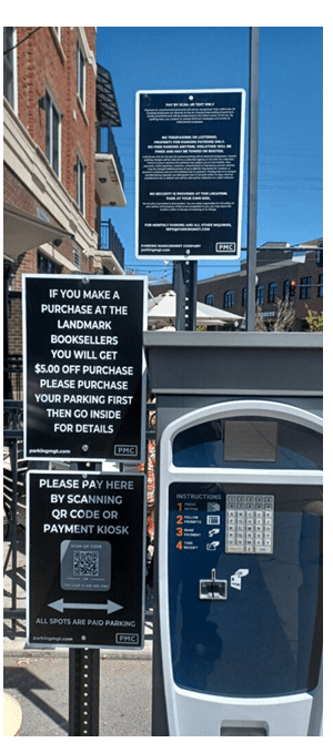 A parking payment kiosk with multiple signs detailing payment instructions, a discount for bookstore purchases, and parking rules, set outdoors on a sunny day in an urban area with buildings in the background.