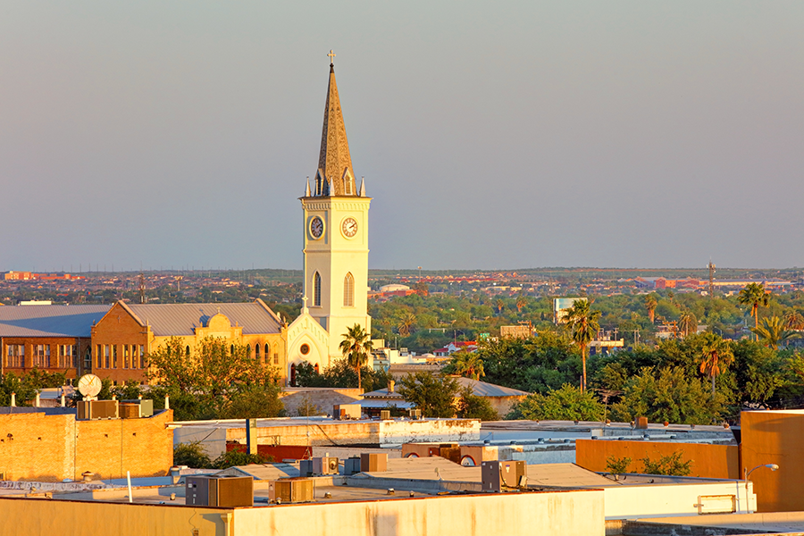A church with a tall steeple and clock stands among trees and buildings at sunset, overlooking a flat landscape under a clear sky.