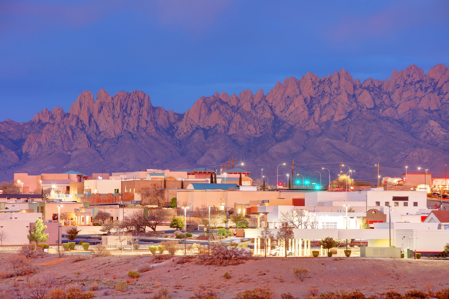 A cityscape at dusk with pastel-colored buildings in the foreground and rugged, rocky mountains in the background under a purple-blue sky.