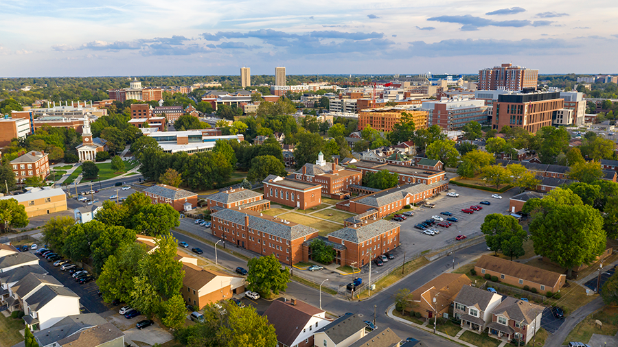 Aerial view of a cityscape with a mix of red-brick buildings, parking lots, leafy trees, and modern high-rise structures under a partly cloudy sky.
