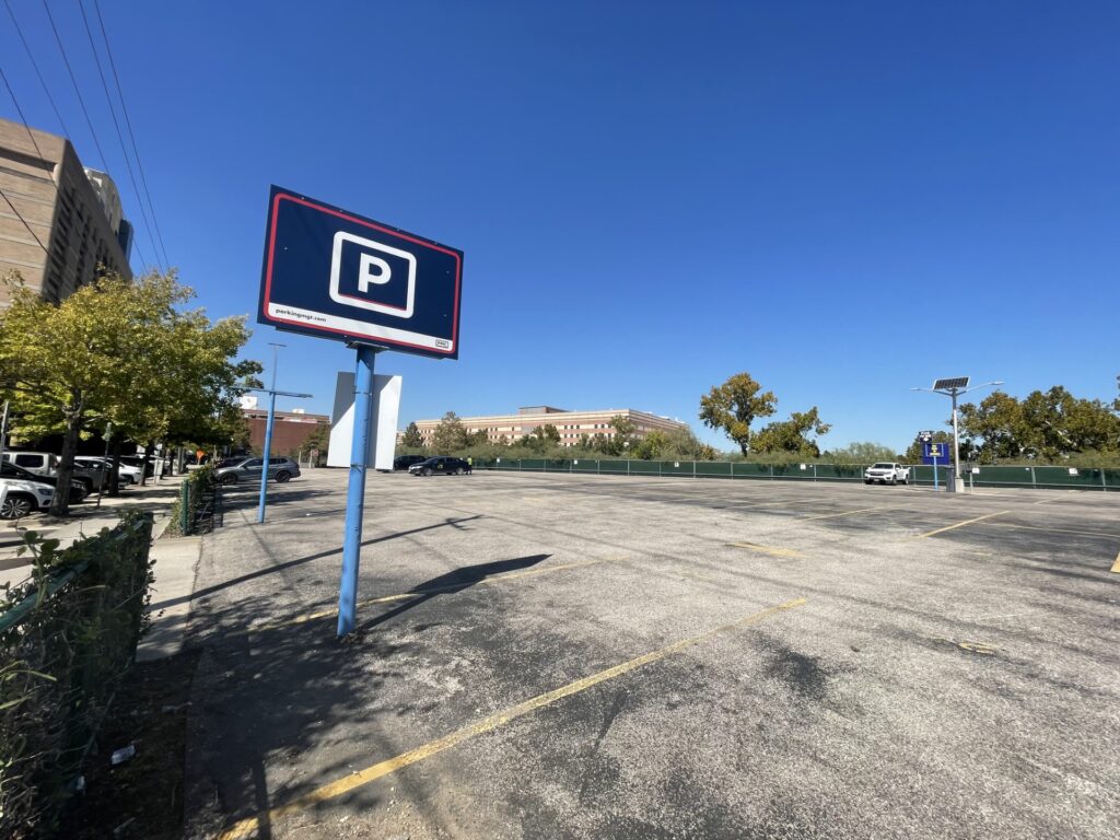 An empty outdoor parking lot with a large blue and white "P" parking sign in the foreground, surrounded by trees and buildings under a clear blue sky.