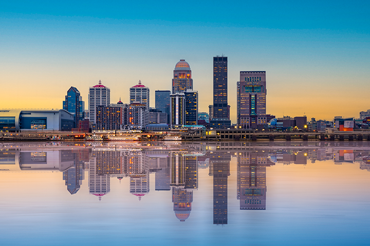 A city skyline with modern high-rise buildings is reflected in calm water at sunset, with a clear sky transitioning from blue to orange.
