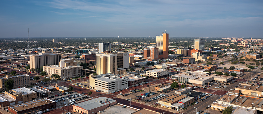 Aerial view of a mid-sized city with several tall office buildings, smaller commercial structures, and wide streets, stretching out to a flat horizon under a partly cloudy sky.
