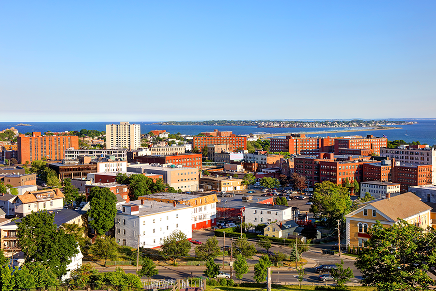 A panoramic view of a coastal city with brick buildings, trees, and roads under a clear blue sky, with the ocean visible in the background.