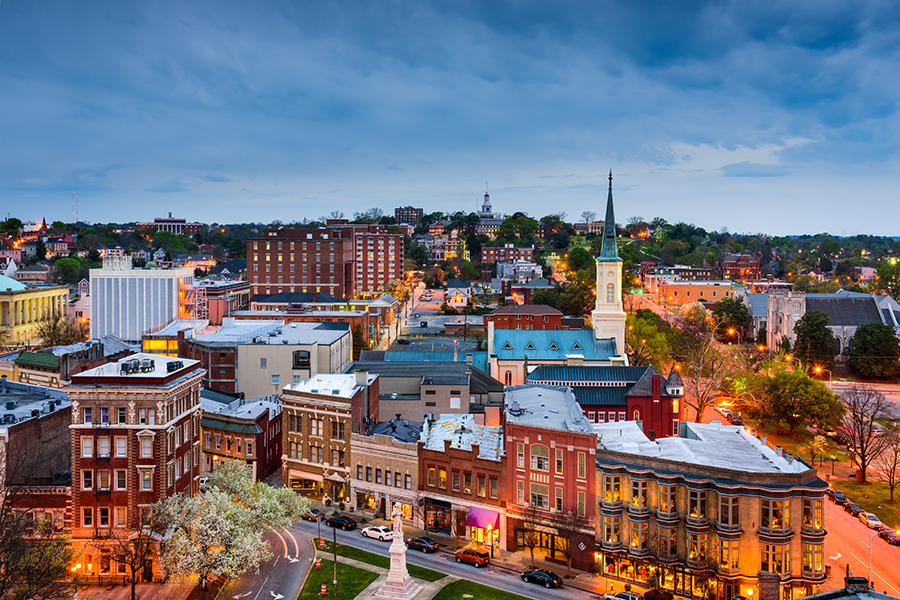 Aerial view of downtown Macon, Georgia, at dusk with historic buildings, colorful facades, a church steeple, and city lights illuminating the streets and trees.