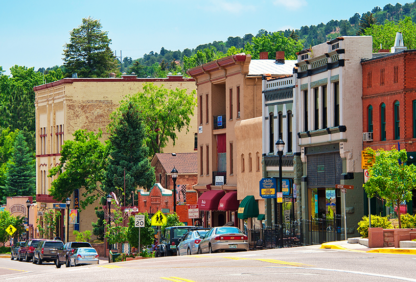 Colorful historic buildings line a tree-filled main street in a small town, with cars parked along the road and green hills visible in the background under a blue sky.