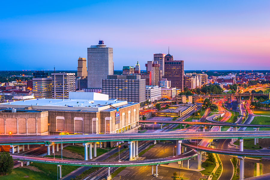 A view of downtown Memphis, Tennessee at sunset, showing tall buildings, a network of highways with blurred car lights, and a pastel-colored sky.
