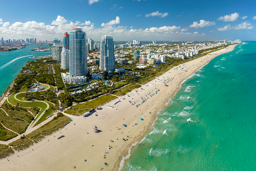Aerial view of a sunny beach lined with turquoise water and white sand, high-rise buildings along the coastline, and a cityscape in the background under a partly cloudy sky.