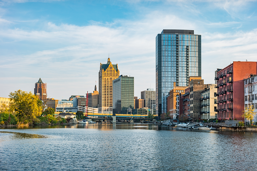 A river runs through a cityscape with tall modern and historic buildings, boats docked along the water, and a clear blue sky above. Trees with yellow-green leaves line the riverbank on the left.