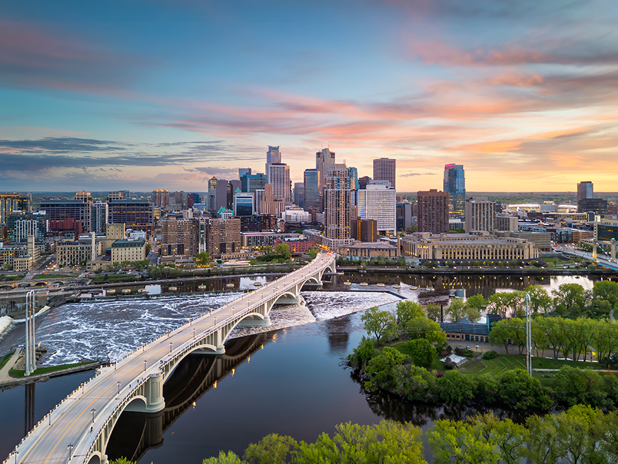 Aerial view of downtown Minneapolis at sunset, featuring a bridge over the Mississippi River, a small waterfall, green trees, and modern skyscrapers under a colorful sky.