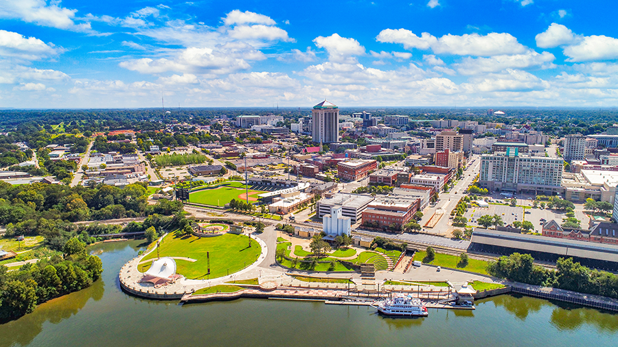 Aerial view of a cityscape with a riverfront park, green spaces, a white amphitheater, downtown buildings, and a boat docked along the waterfront under a partly cloudy blue sky.