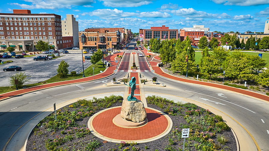 A roundabout with a statue on a rock in the center, surrounded by landscaped greenery, leads into a downtown area with brick buildings, parked cars, and a partly cloudy sky overhead.