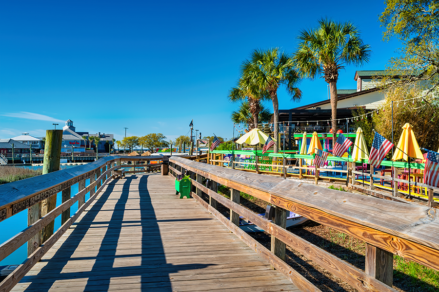 A wooden boardwalk with railings runs alongside a colorful outdoor dining area with umbrellas and American flags. Palm trees and clear blue skies complete the sunny, vibrant scene.