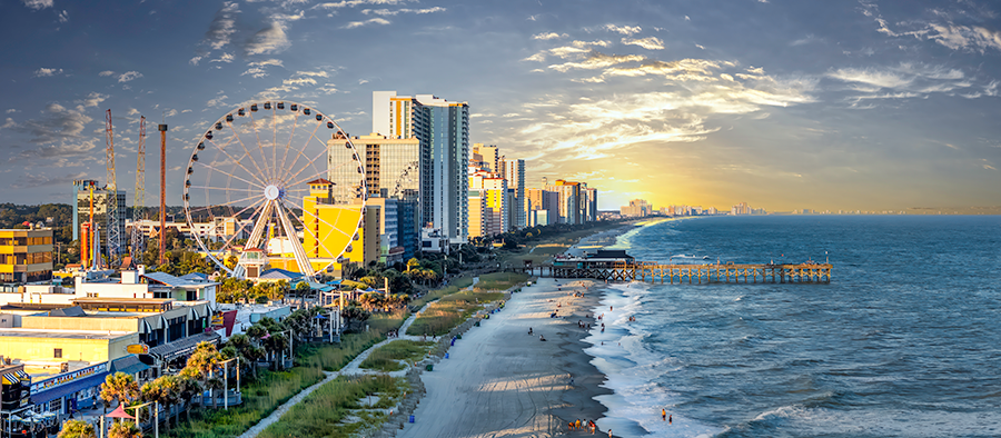 A vibrant city beachscape at sunset features tall hotels, a large Ferris wheel, an amusement park, and a long pier extending into the ocean, with waves gently reaching the sandy shore.
