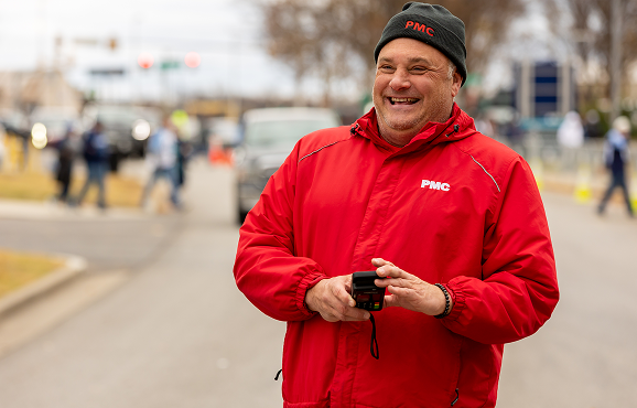 A smiling man in a red PMC jacket stands outdoors on a street, holding a small device. There are blurred people, cars, and trees in the background.
