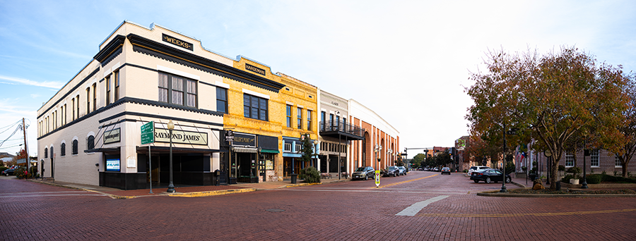 A wide view of a quiet downtown street with historic brick buildings, shops, and trees with autumn leaves; the street is paved with red bricks and a few cars are parked along the sidewalks.