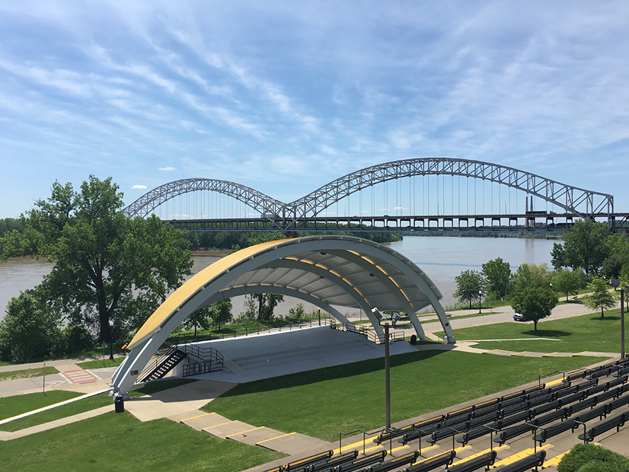 An outdoor amphitheater with a curved white roof sits near a river, with a large arched steel bridge spanning the water in the background under a partly cloudy sky. Rows of seating and green lawns surround the stage.