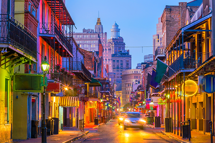 Colorful buildings with balconies line a lively street in New Orleans’ French Quarter at dusk, with glowing lights and blurred cars, and tall city buildings in the background.