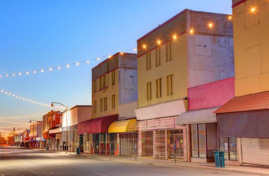 A quiet, empty street lined with colorful, old storefronts and string lights overhead at dusk, with closed shops and a clear blue sky.