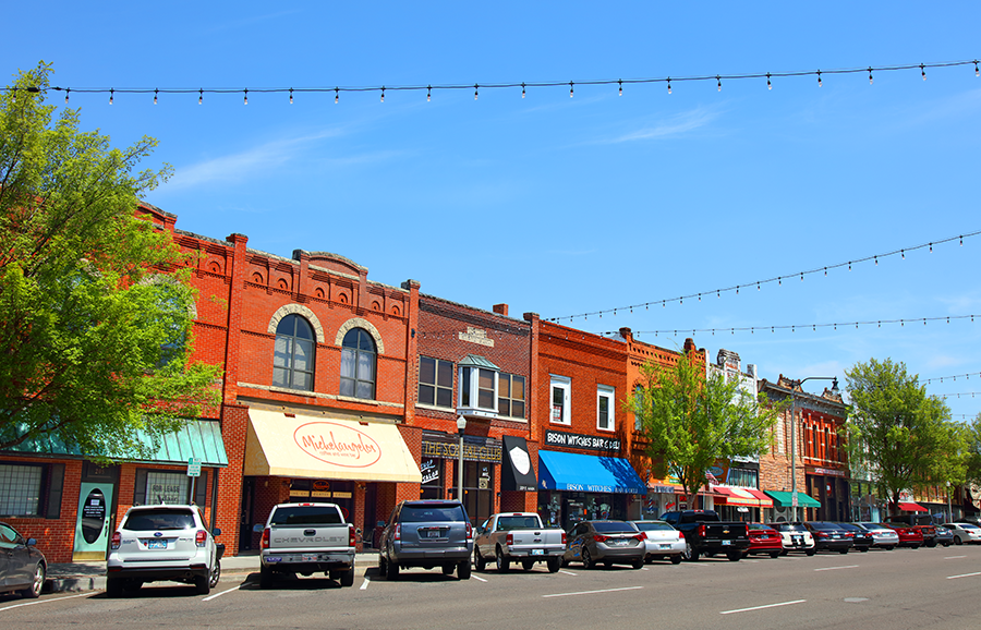 A row of colorful brick storefronts lines a small-town street under a clear blue sky, with parked cars along the roadside and string lights hanging overhead.
