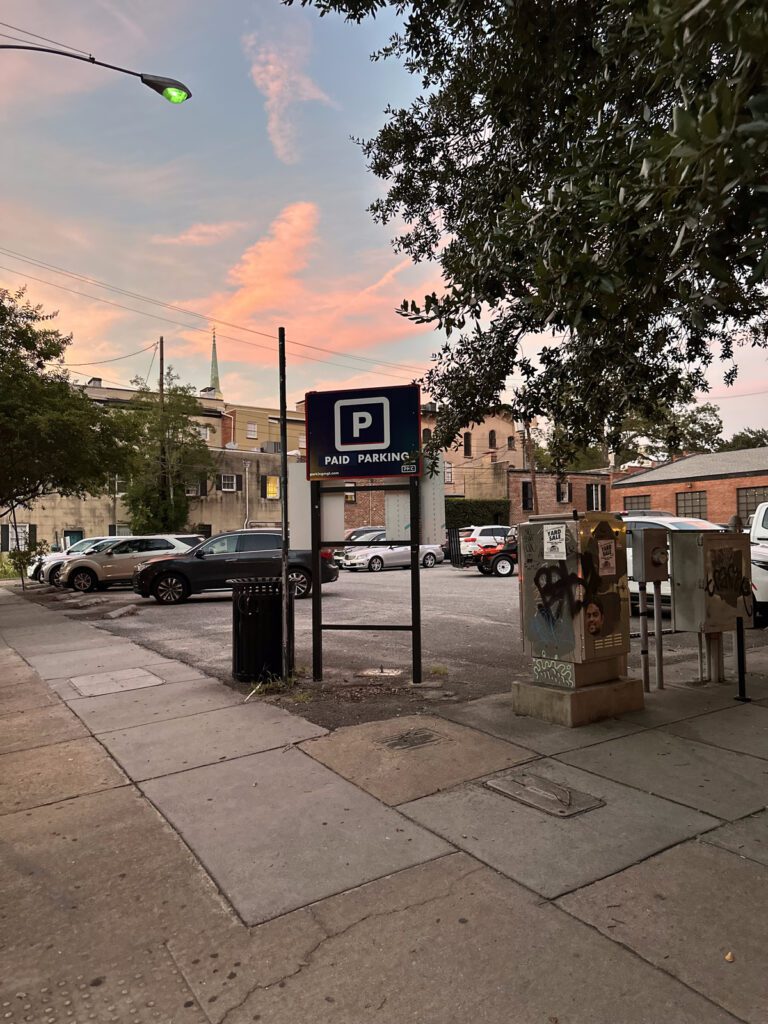 A sidewalk at sunset with a PMC "Paid Parking" sign in front of a parking lot filled with cars. Trees, utility poles, and nearby buildings are visible in the background.