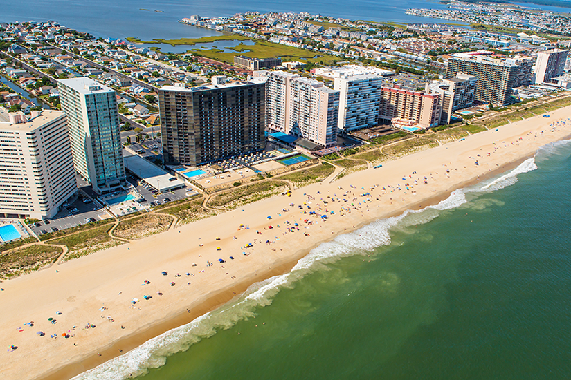 Aerial view of a beach lined with high-rise hotels and condominiums, calm waves, and people scattered along the sandy shore. Residential neighborhoods and waterways are visible in the background.