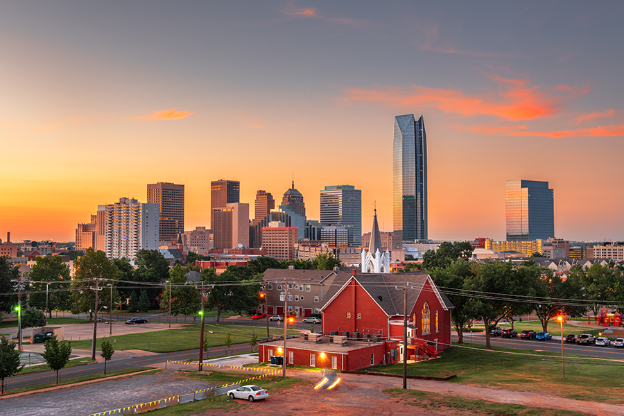 Oklahoma City skyline at sunset with modern skyscrapers, a historic red brick church in the foreground, and vibrant orange and blue hues in the sky. Trees and parked cars surround the buildings.