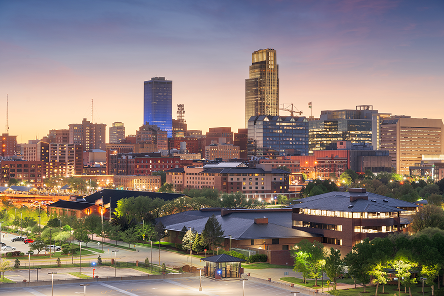 A city skyline at sunset with modern tall buildings, shorter brick structures, and greenery in the foreground. The sky has soft pastel hues, and city lights are starting to illuminate the scene.