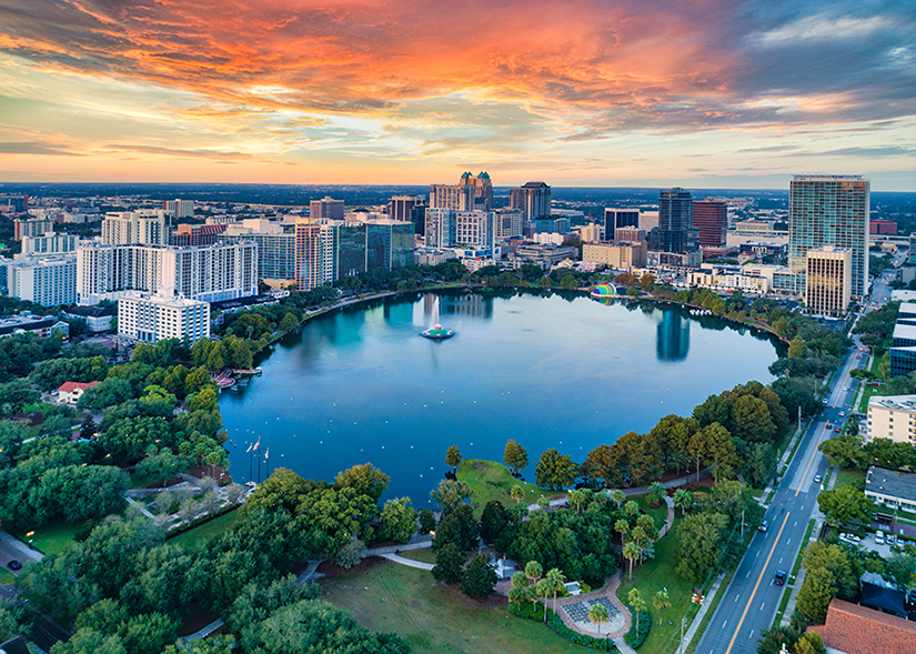 Aerial view of a city skyline at sunset, featuring a large circular lake surrounded by green parks, trees, and tall modern buildings under a colorful sky.