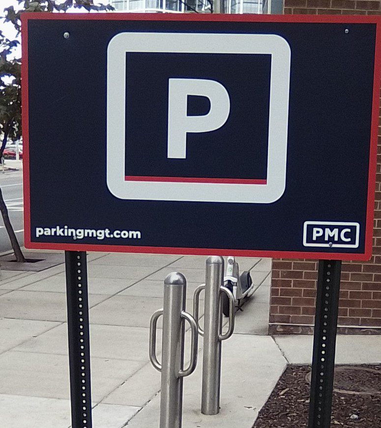 A parking sign with a large white "P" on a dark background, displaying the website "parkingmgt.com" and the logo "PMC," is posted on a sidewalk near a building and bike racks.