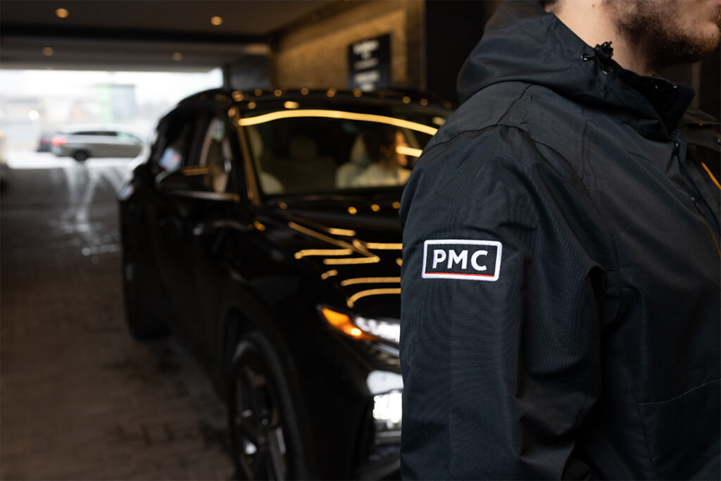 A person wearing a black jacket with a "PMC" patch on the sleeve stands near a black car in a covered driveway. The car’s headlights and reflections are visible.
