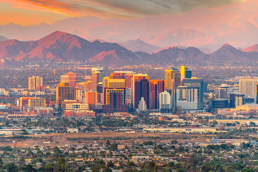 Downtown Phoenix skyline at sunset, with tall modern buildings in the foreground and rugged mountains in the background under a colorful sky.