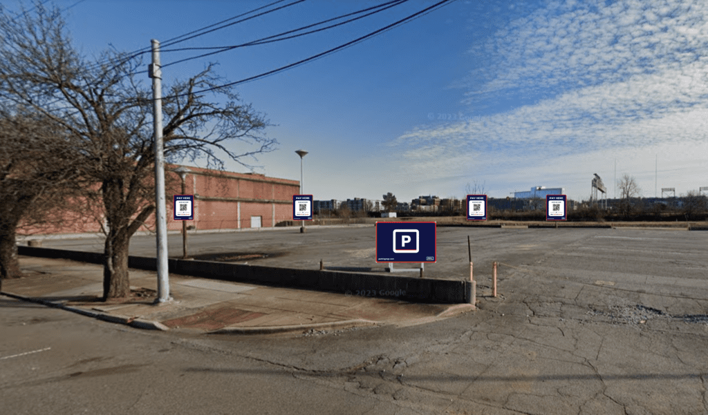 An empty, cracked asphalt parking lot with several parking signs, surrounded by a low curb, a bare tree, a utility pole, and a red brick building on the left under a partly cloudy sky.