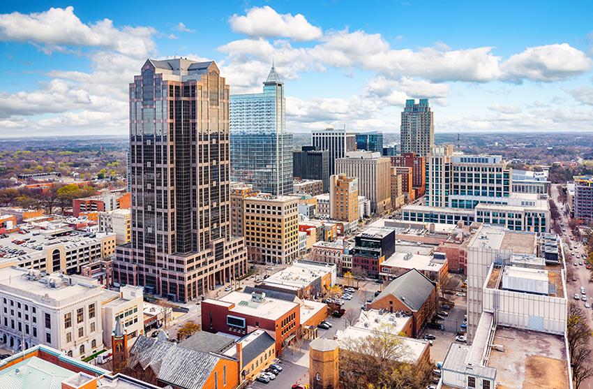 Aerial view of downtown Raleigh, North Carolina, featuring modern skyscrapers, mid-rise buildings, and tree-lined streets under a partly cloudy blue sky.