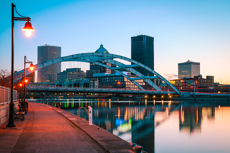 A cityscape at dusk featuring a large arched bridge over a calm river, with reflections of city buildings and street lamps lining a riverside walkway. The sky is clear with a soft gradient from blue to orange.
