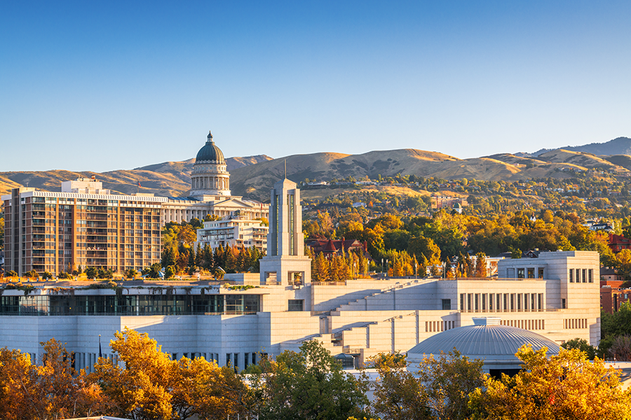 Cityscape of Salt Lake City, Utah, with the Utah State Capitol building, modern structures, trees with autumn foliage, and mountains in the background under a clear blue sky.