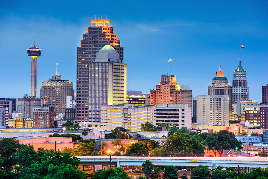 San Antonio downtown skyline at dusk, featuring lit-up high-rise buildings, the Tower Life Building, and the illuminated Tower of the Americas against a blue evening sky. Trees and highways are visible in the foreground.