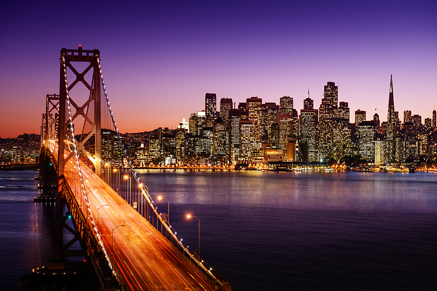 The San Francisco skyline at dusk with city lights shining, viewed from behind the illuminated Bay Bridge stretching over the water. The sky is a gradient of deep blue and purple.