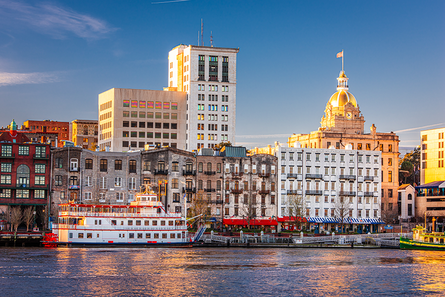 Historic buildings and a riverboat line the riverfront in Savannah, Georgia, with the sun casting a warm glow on the architecture, including a domed building, under a clear blue sky.