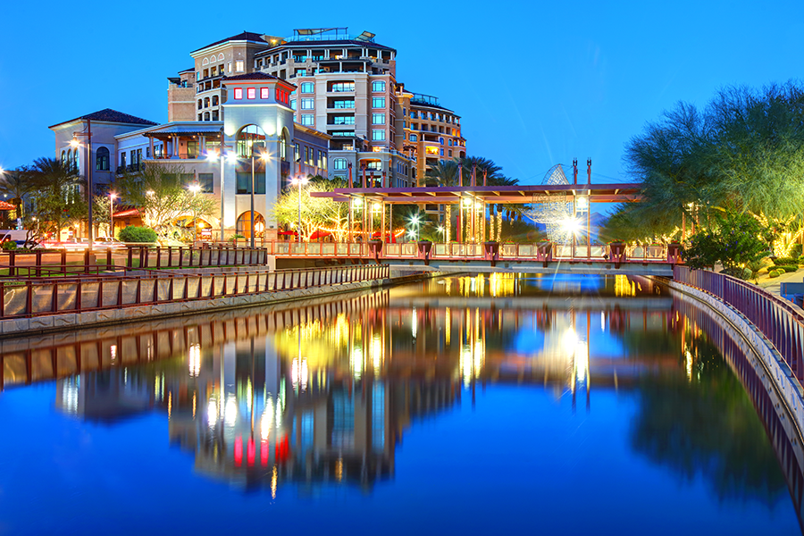Modern buildings and palm trees are reflected in a calm river at dusk in Scottsdale, Arizona. The sky is deep blue, and bright lights from the structures and bridge create colorful reflections on the water.