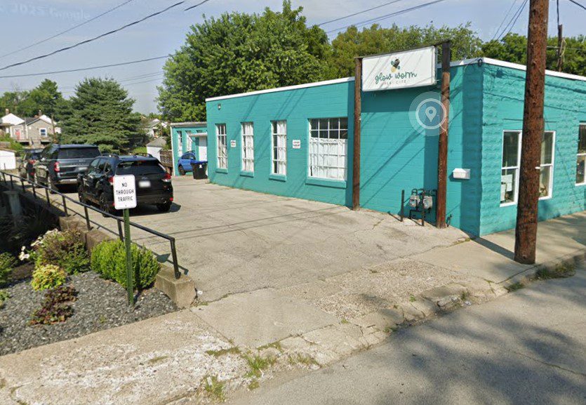 A turquoise one-story building with large windows sits next to a small parking lot. Several cars are parked outside, and a "No Thru Traffic" sign is posted near a landscaped area with shrubs.